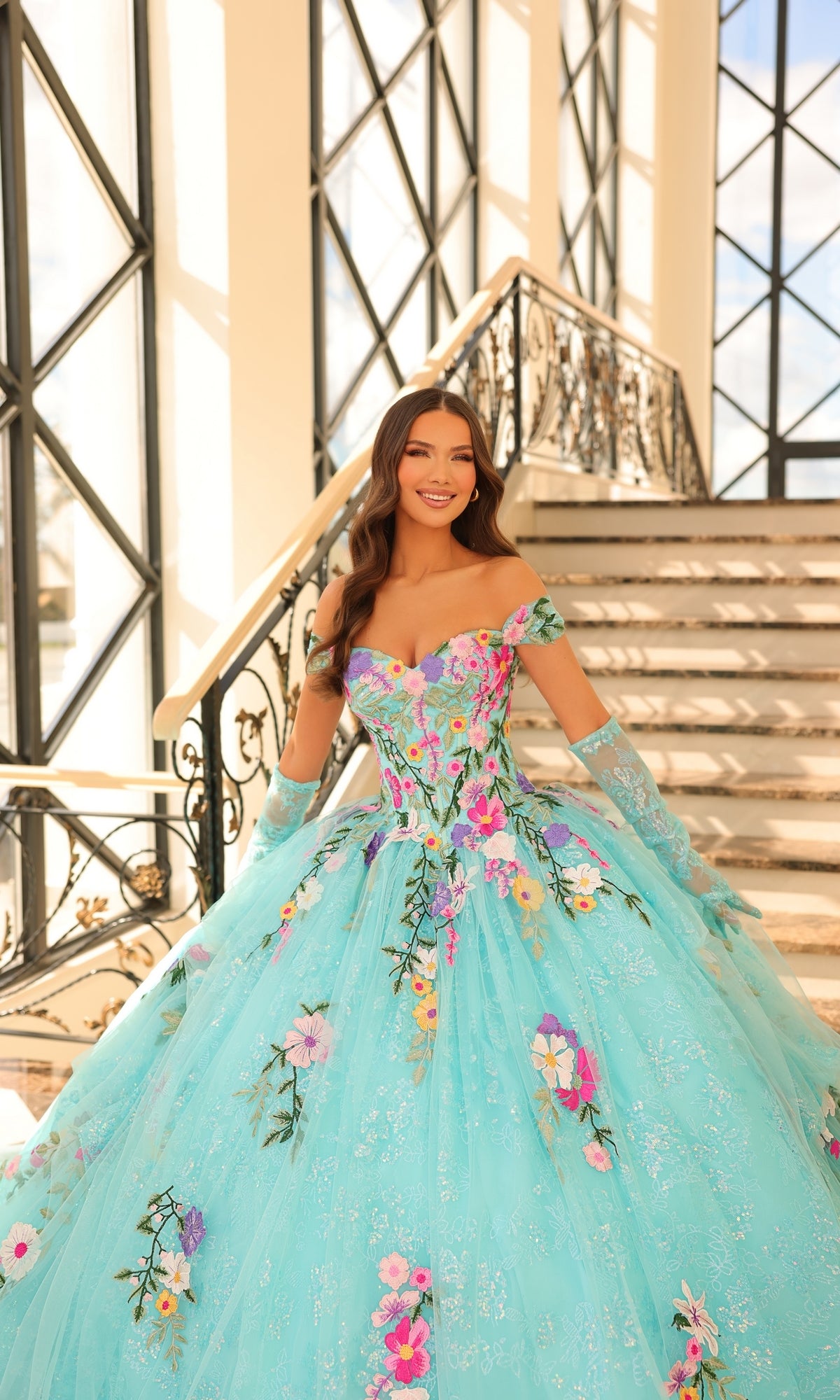 A woman wearing the Quinceanera Dress 54200 by Amarra, an off-the-shoulder gown with floral embroidery, stands at the base of a staircase in a sunlit, elegant interior.